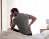 Man sat on bed facing away from camera holding his lower back in pain with lead in hand.