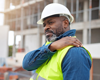 Construction man in fifties holding shoulder in pain. Wearing hat and work clothes.