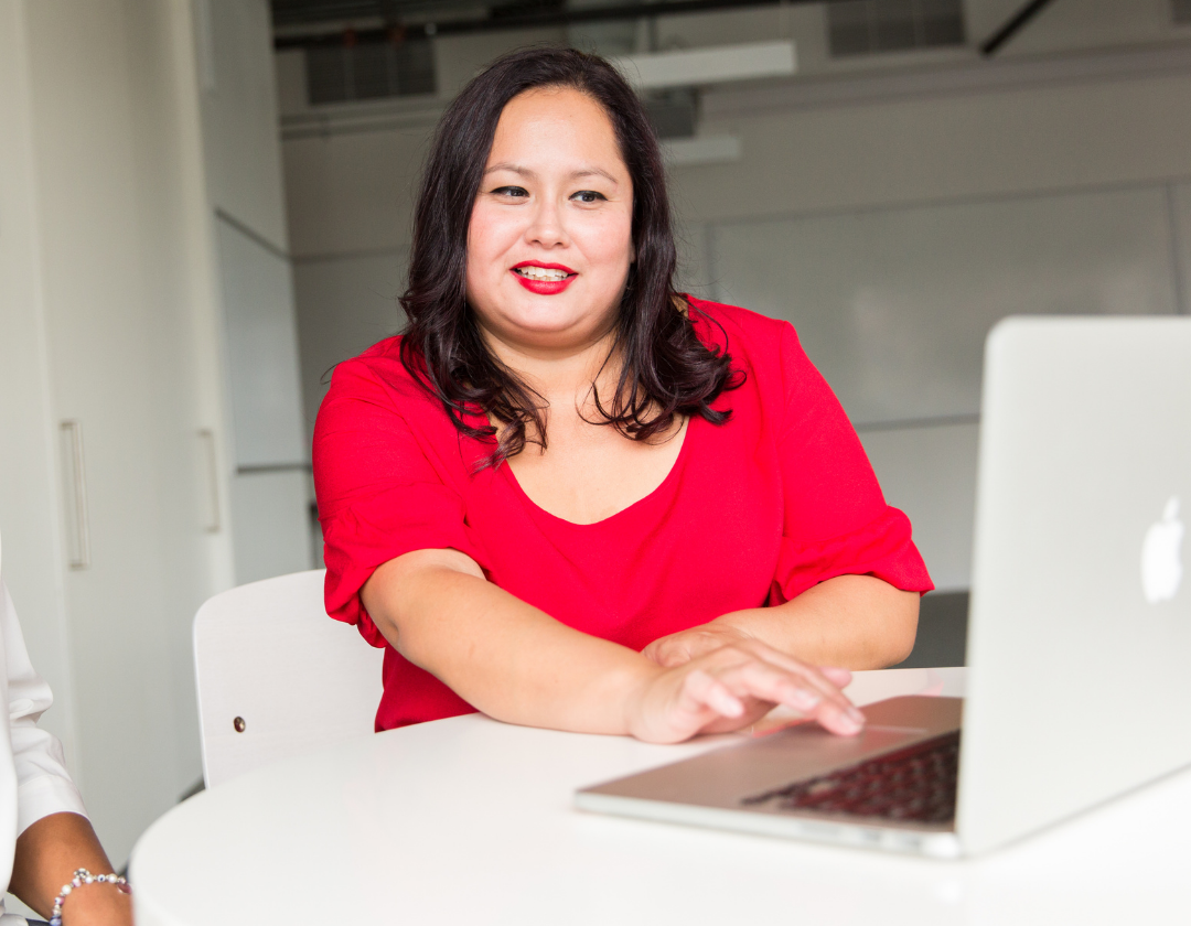 Woman in red top and lipstick sat by computer smiling