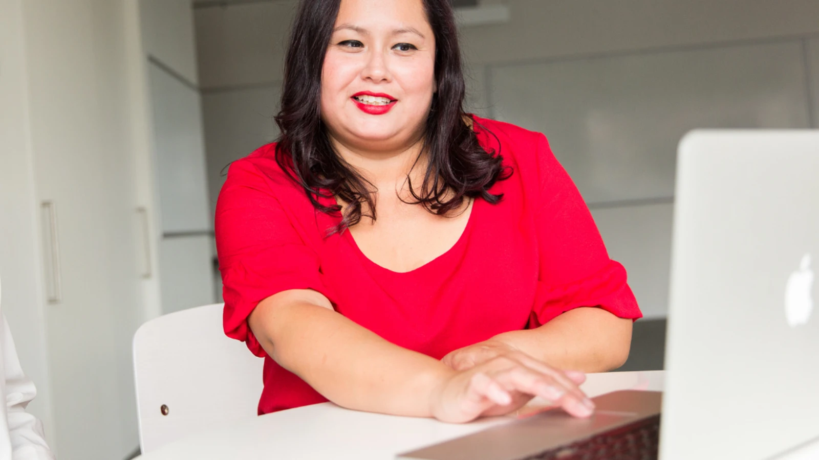 Woman in red top and lipstick sat by computer smiling