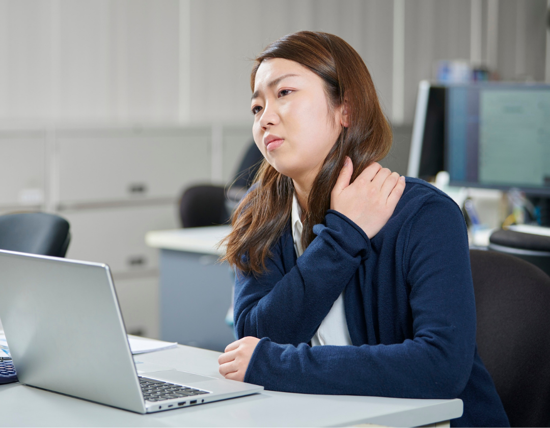 Young woman looking sad, sat at office desk holding neck in pain.